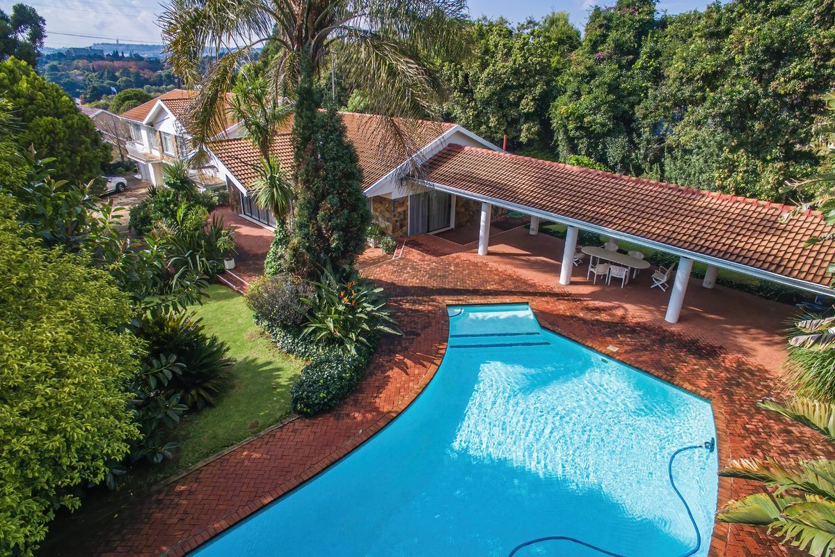 Aerial view of a residential property featuring a large swimming pool, extensive brick-paved patio area, covered walkway, and mature landscaped gardens.