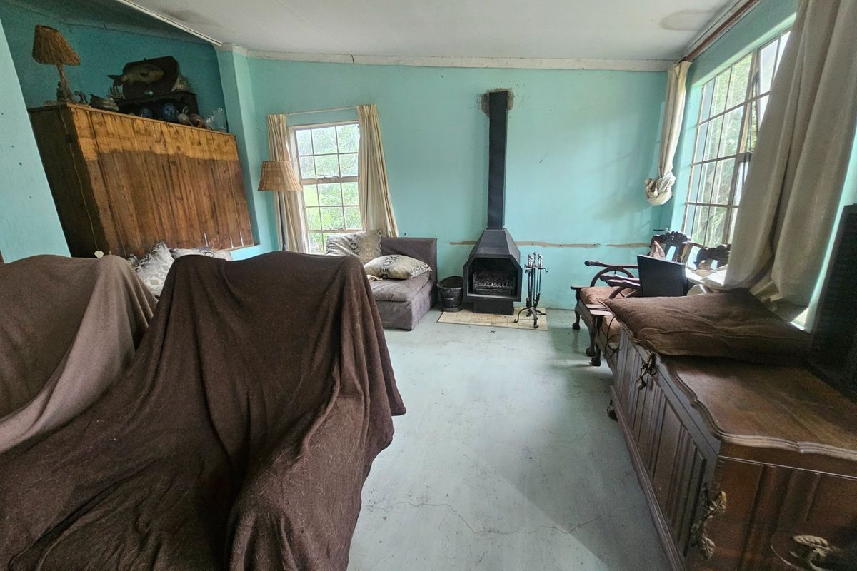 A living room featuring a freestanding fireplace, timber-framed windows, and painted concrete flooring, requiring significant cosmetic updates and repairs to the ceiling and walls.