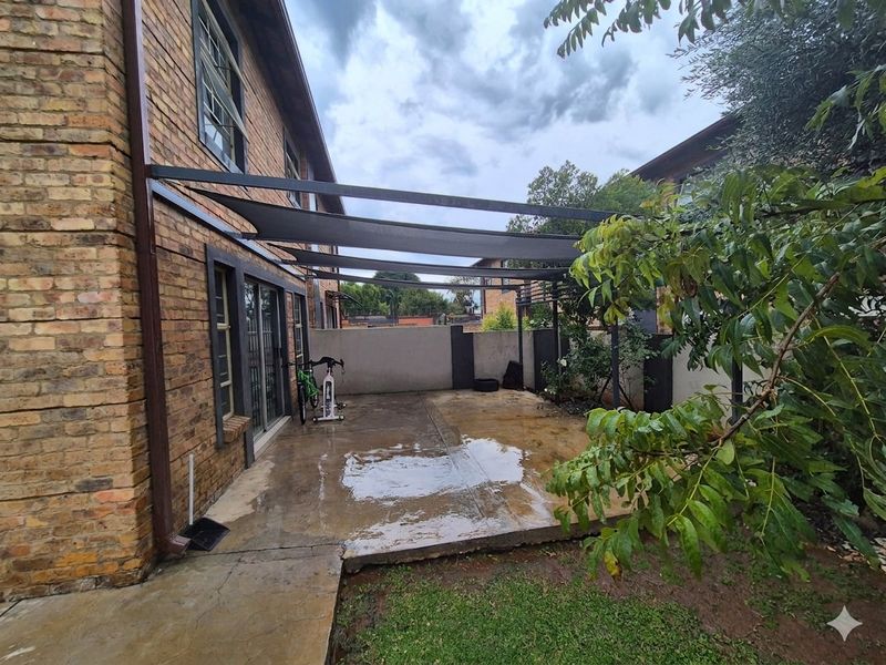 A paved rear patio area featuring a shade sail structure, brick exterior walls, and direct access to the interior via sliding glass doors.