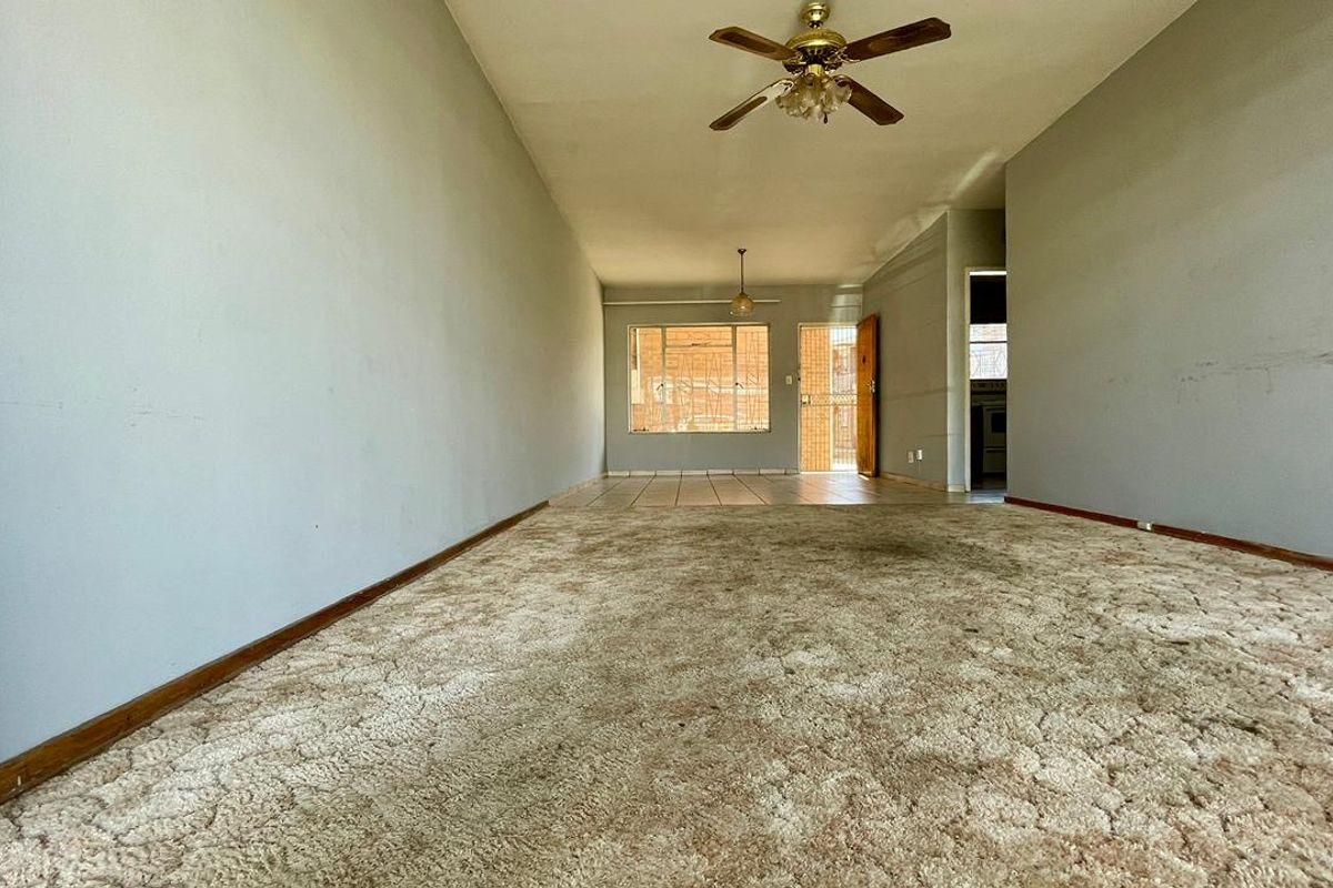 Spacious, empty living area featuring a ceiling fan, carpeted flooring, and access to a secure entrance with burglar bars.