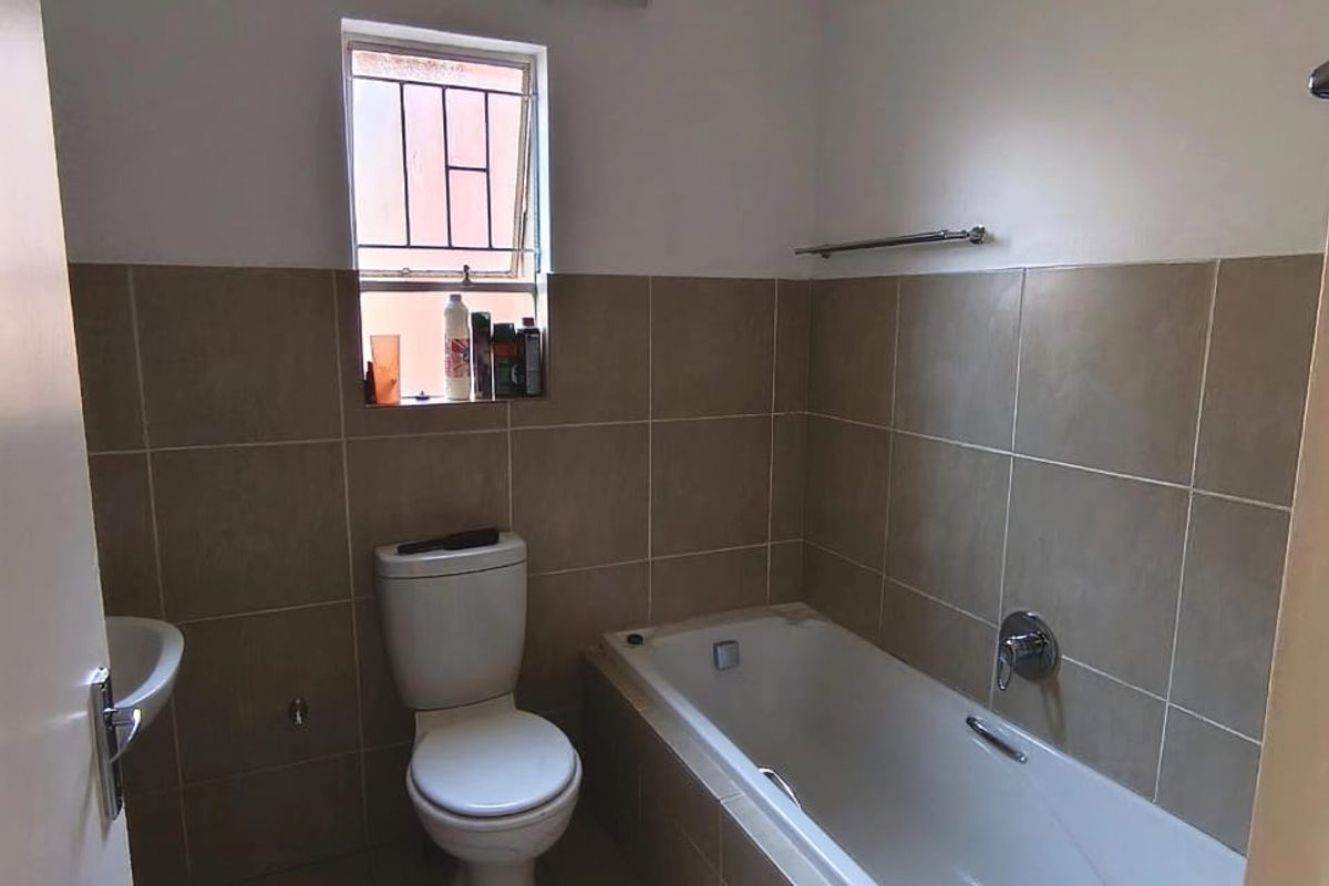 A functional bathroom featuring a bathtub, toilet, and wall-mounted basin with neutral tiled finishes and a window with security bars.