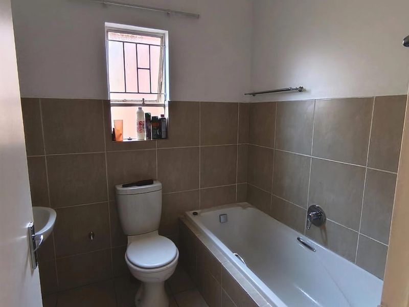 A functional bathroom featuring a bathtub, toilet, and wall-mounted basin with neutral tiled finishes and a window with security bars.