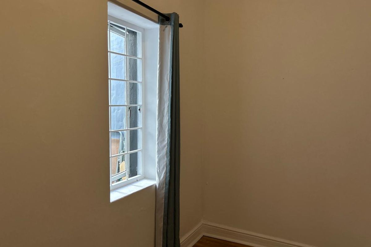 A simple, empty bedroom corner featuring timber flooring, white walls, and a window fitted with security bars.