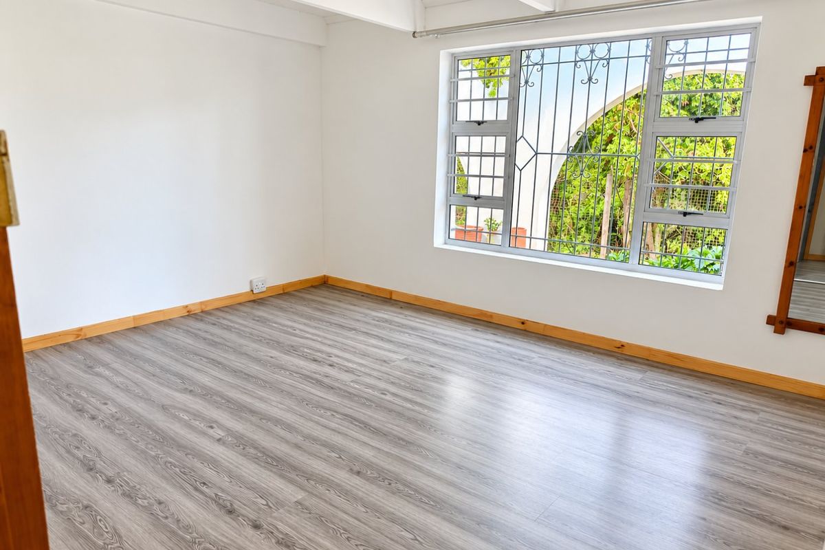 A bright, empty bedroom featuring modern grey laminate flooring, timber skirting boards, and a window fitted with security bars.
