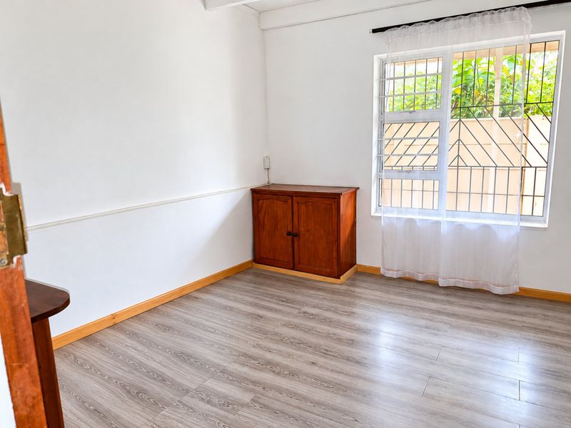 A bright, empty bedroom featuring light-toned laminate flooring, white walls with a chair rail, and a window fitted with security bars.