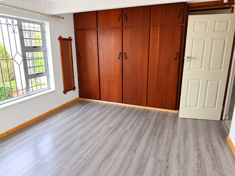 A bright, empty bedroom featuring modern grey laminate flooring, built-in wooden cupboards, and a window fitted with security bars.