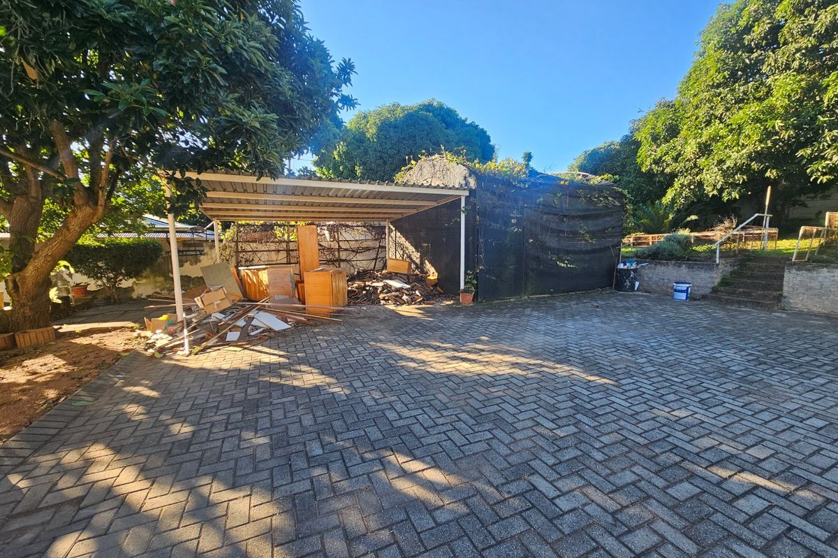 Rear exterior area featuring a paved driveway, a carport structure, and stone steps leading to an elevated garden section.