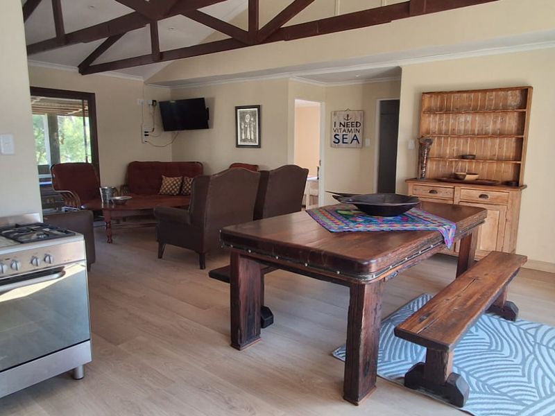Open-plan living and dining area featuring exposed timber roof trusses, light laminate flooring, and a functional kitchen space with a freestanding gas stove.
