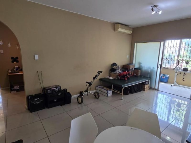 Open-plan living area featuring tiled flooring, a wall-mounted air conditioner, and an arched entryway leading to the kitchen, with access to a balcony.