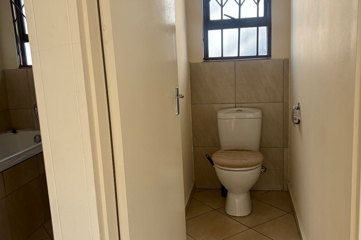 A functional bathroom featuring a toilet, tiled flooring, and a window with security bars, with an adjacent view of a bathtub.
