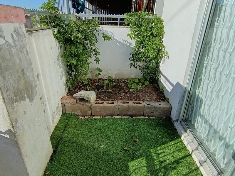 A small outdoor patio area featuring artificial grass, a concrete block planter, and a covered roof structure, providing a compact space for outdoor use.