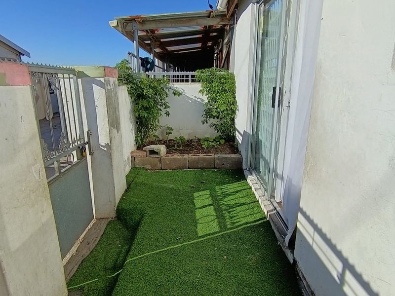 A narrow outdoor patio area featuring artificial turf, a sliding glass door entrance, and a metal gate, showing signs of wear on the exterior walls.