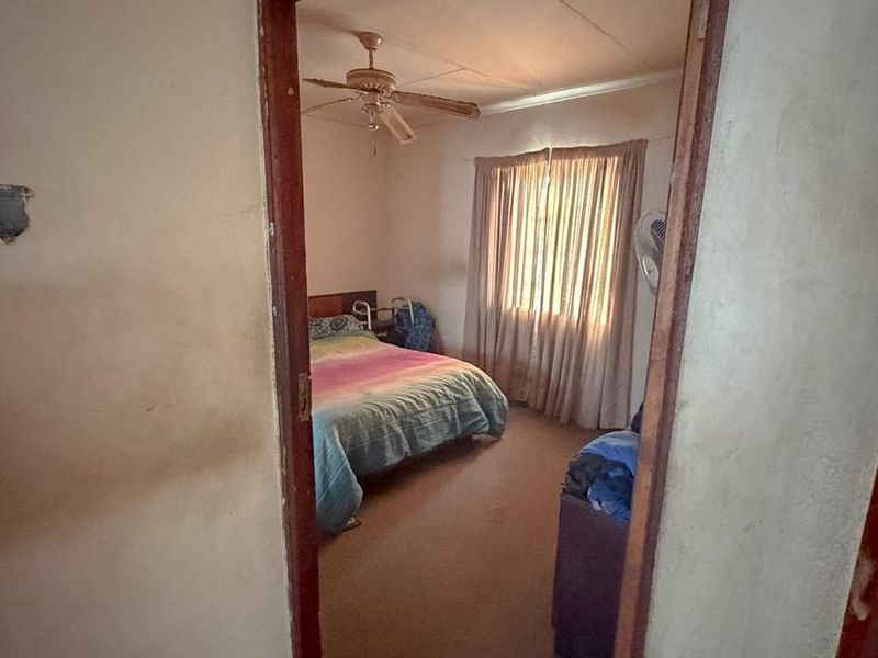 A bedroom viewed through a doorway, featuring a ceiling fan and window with curtains. The room shows signs of wear and requires cosmetic updates.