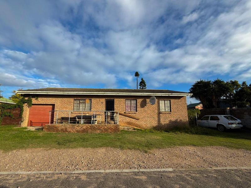 Single-story face-brick house featuring a raised front patio, integrated garage, and tiled roof, situated on a large plot with a gravel driveway.