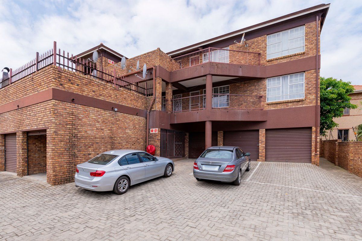 Front exterior view of a multi-story brick apartment building featuring paved parking, garage access, and private balconies with metal railings.