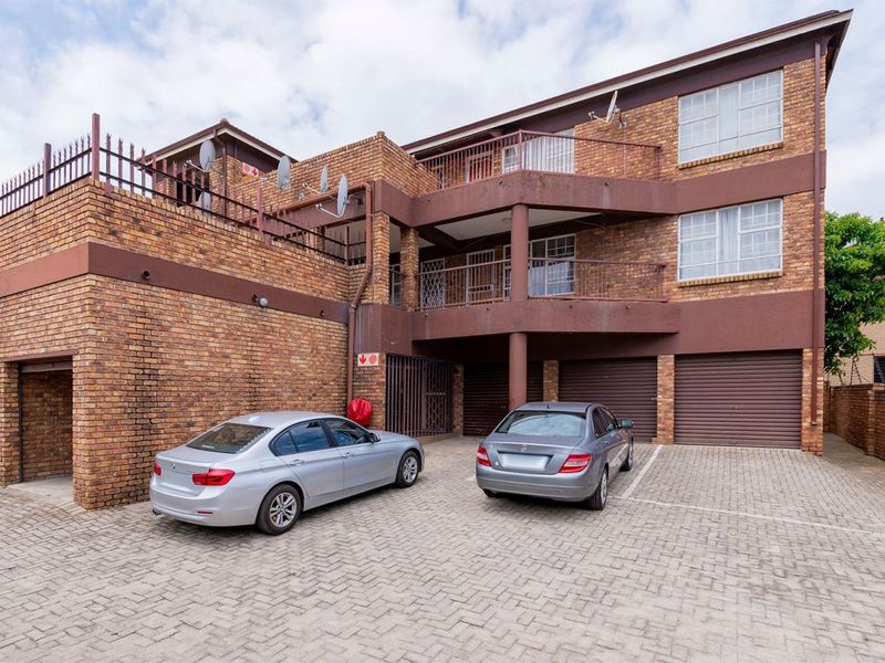 Front exterior view of a multi-story brick apartment building featuring paved parking, garage access, and private balconies with metal railings.