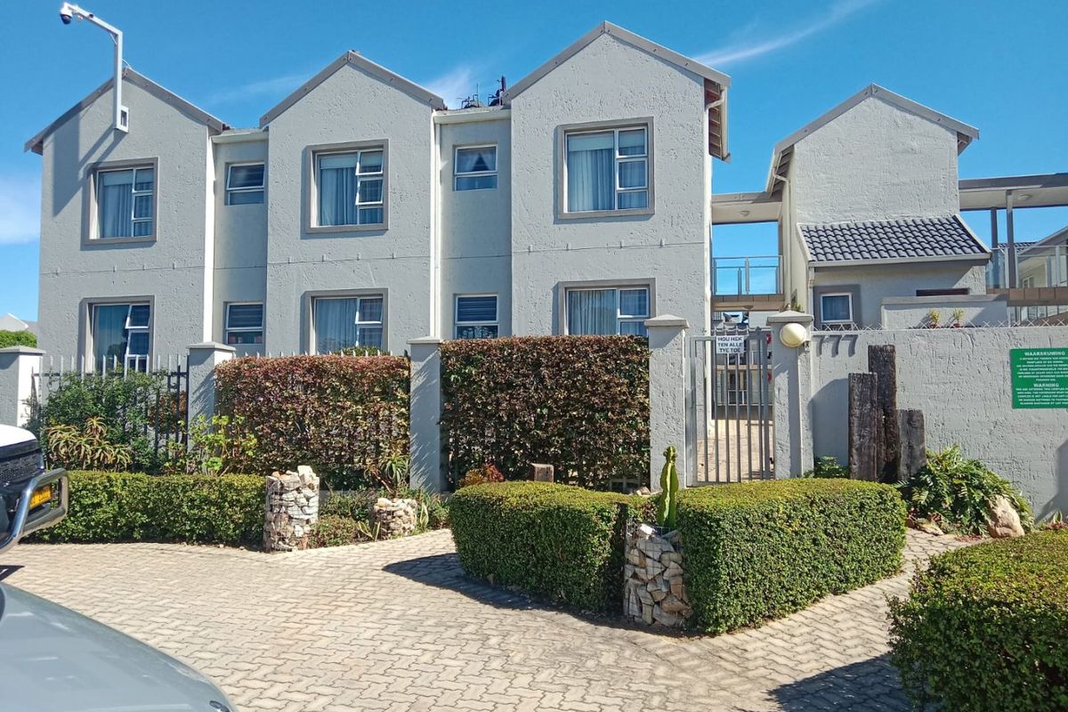 Front elevation of a multi-story apartment complex featuring a paved driveway, secure gated entrance, and well-maintained landscaped hedging.