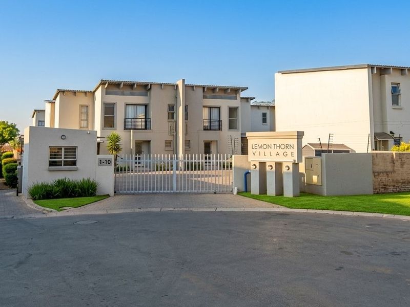 Front entrance view of Lemon Thorn Village apartment complex featuring a secure gated access point, paved driveway, and perimeter wall with electric fencing.