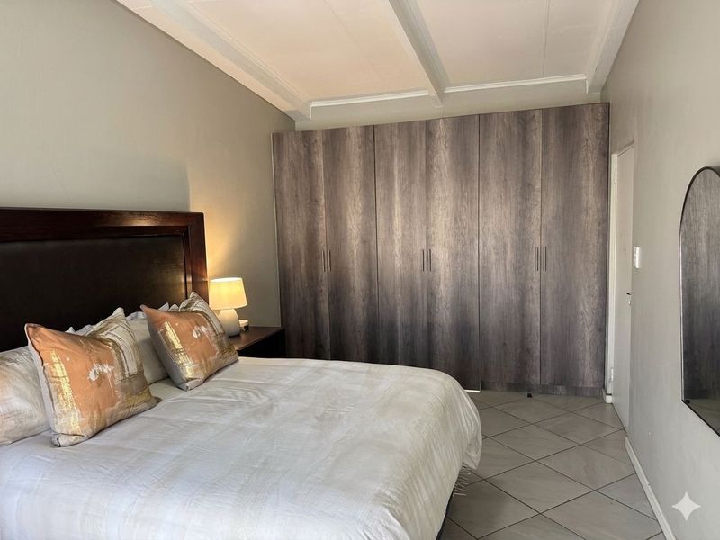 A well-maintained bedroom featuring extensive built-in wooden cupboards, neutral tiled flooring, and exposed ceiling beams, providing a clean and functional living space.