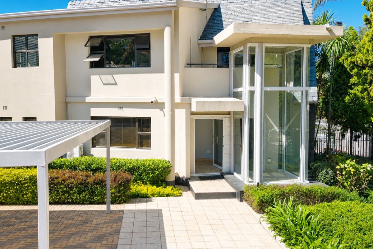 Rear exterior view of a commercial property featuring a modern glass elevator shaft, paved patio area, metal pergola, and well-maintained landscaped garden.