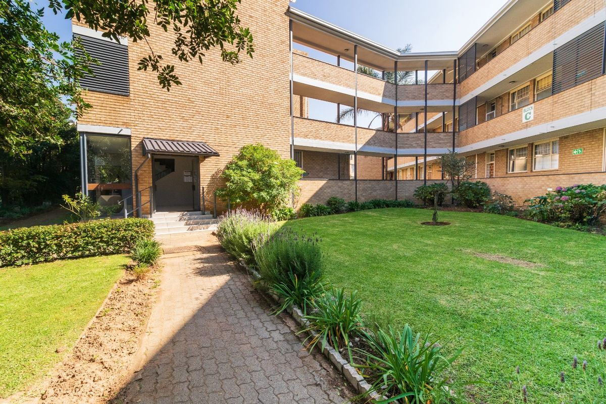 A well-maintained apartment complex exterior featuring a brick facade, paved walkway, and landscaped garden area with multi-level balcony access.