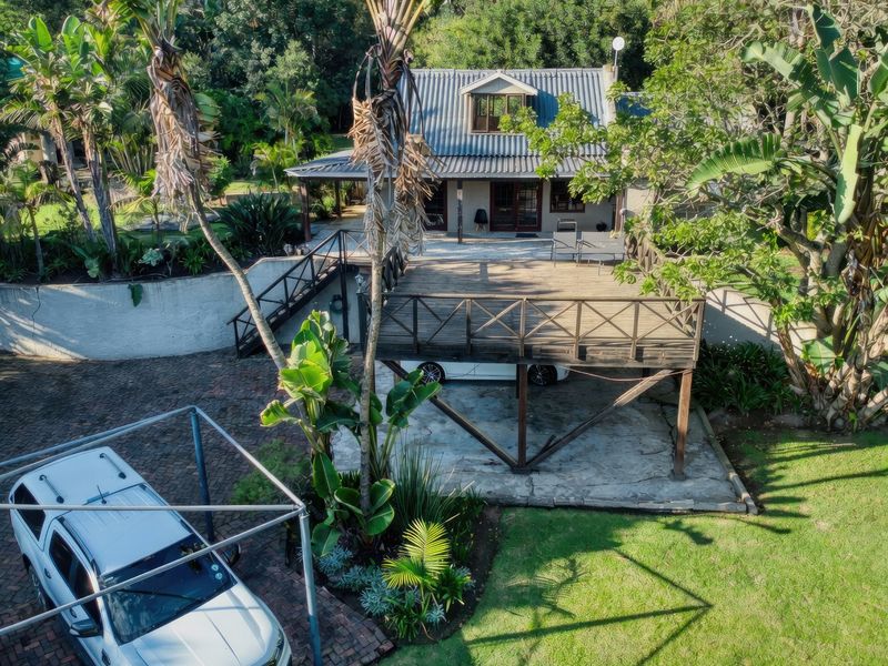 Aerial view of a farm property featuring a raised timber deck, paved driveway, carport, and a main dwelling with a corrugated metal roof.