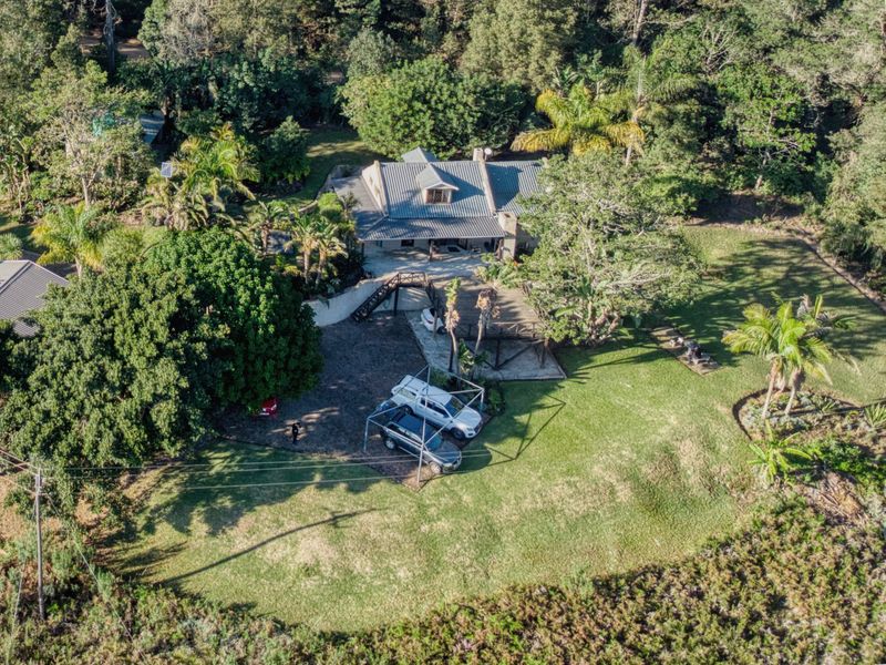 Aerial view of a farm property featuring a main dwelling with a metal roof, expansive lawn, mature trees, and a timber deck.