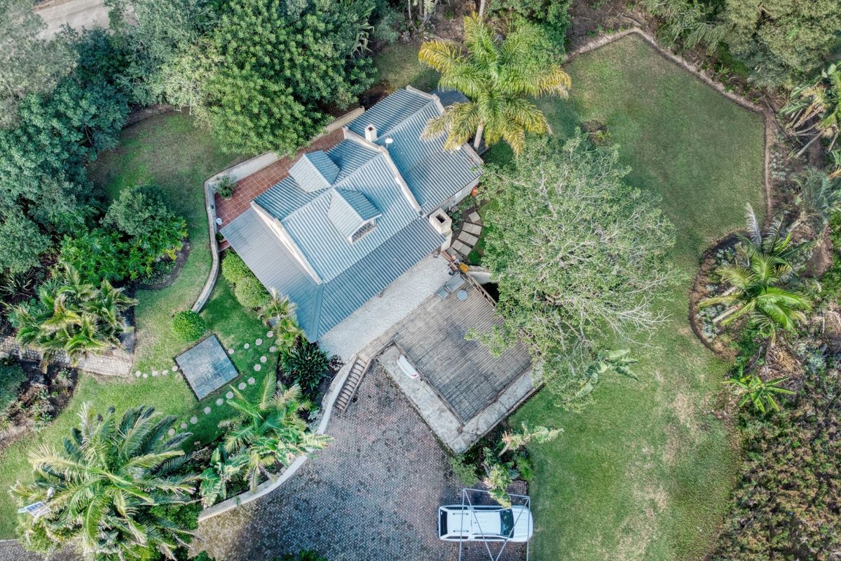 Aerial view of a farm property featuring a metal-roofed residence, expansive landscaped gardens, a timber deck, and a paved driveway area.