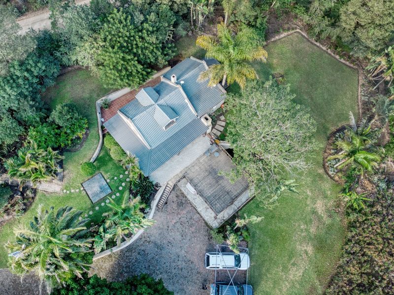 Aerial view of a farm property featuring a metal-roofed residence, expansive landscaped gardens, a timber deck, and a paved driveway area.