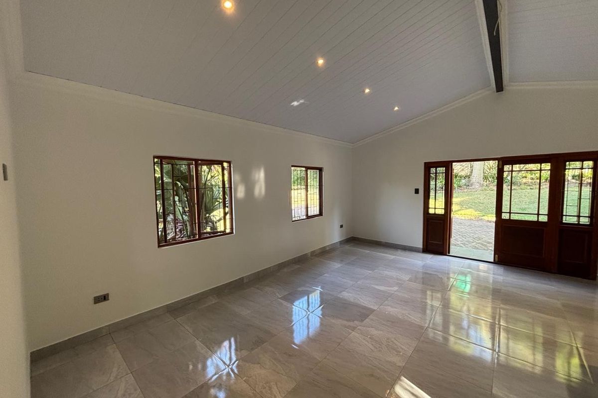 A bright, empty living room featuring polished tiled floors, a vaulted ceiling with recessed lighting, and timber-framed doors opening to the garden.