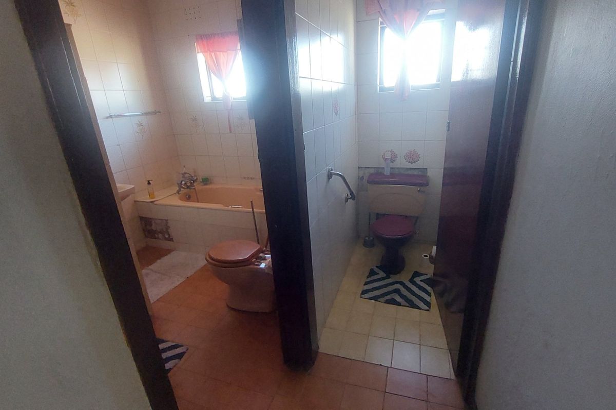 A dated bathroom featuring a separate toilet area, bathtub, and tiled finishes, currently showing signs of wear and missing wall tiles.