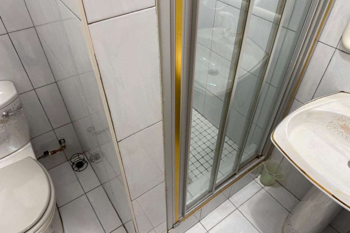 A compact bathroom featuring a pedestal sink, a toilet, and a glass-enclosed shower stall with dated tile and gold-tone fixtures.