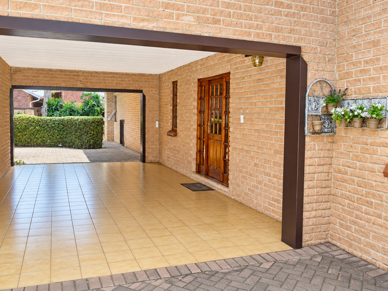 A covered patio or carport area featuring brick walls, tiled flooring, and a timber-framed entrance door, providing a functional outdoor transition space.