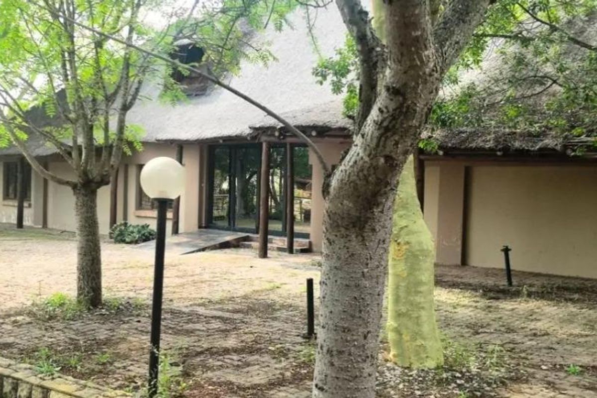 Exterior view of a farm-style dwelling featuring a traditional thatched roof, paved courtyard area, and mature trees surrounding the property.