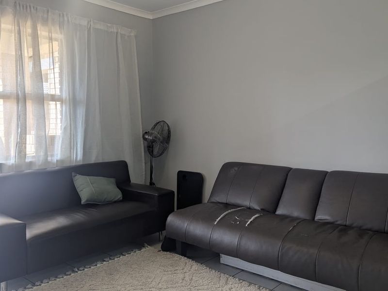 A living room area featuring neutral wall paint, tiled flooring, and a window with sheer curtains.