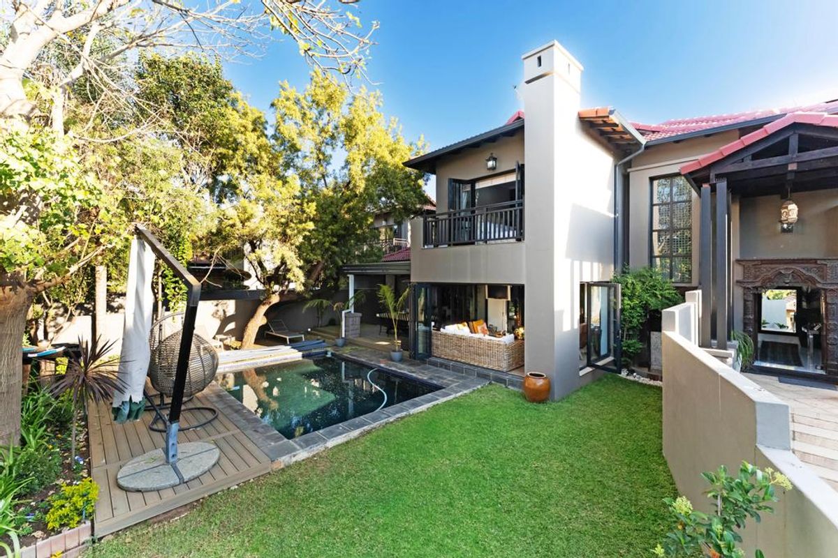 Rear exterior view featuring a swimming pool, timber decking, landscaped garden, and a two-story home with a balcony and bi-fold doors.