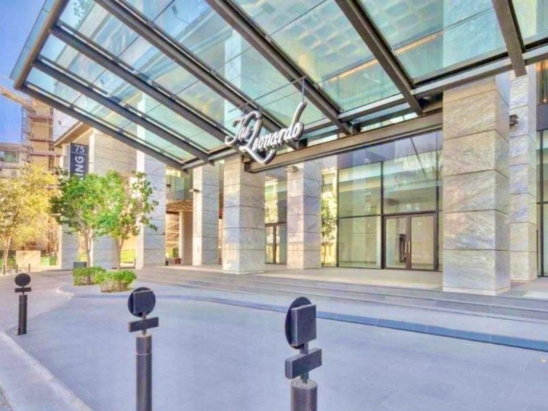 Modern apartment building entrance featuring a prominent glass canopy, stone-clad pillars, and a clean, paved driveway approach.