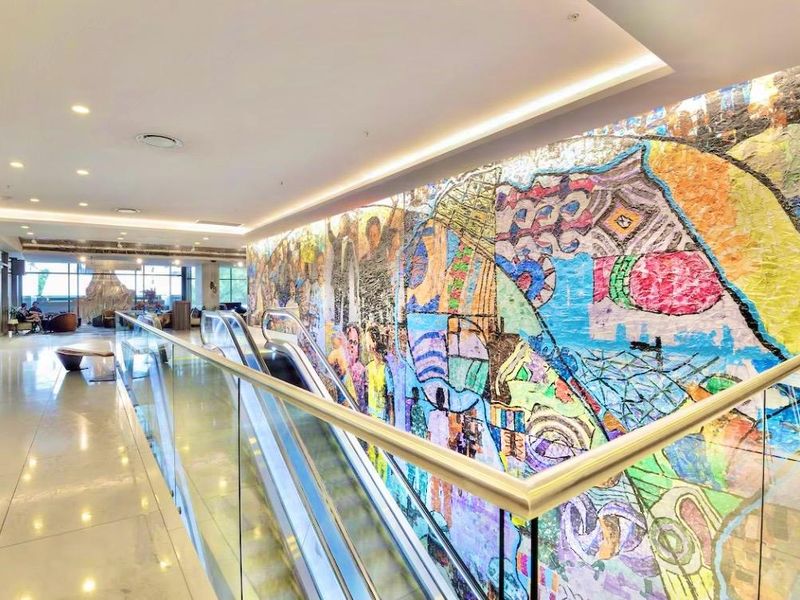 A modern, well-lit hallway featuring polished tile floors, a glass balustrade overlooking an escalator, and a prominent decorative wall mural.