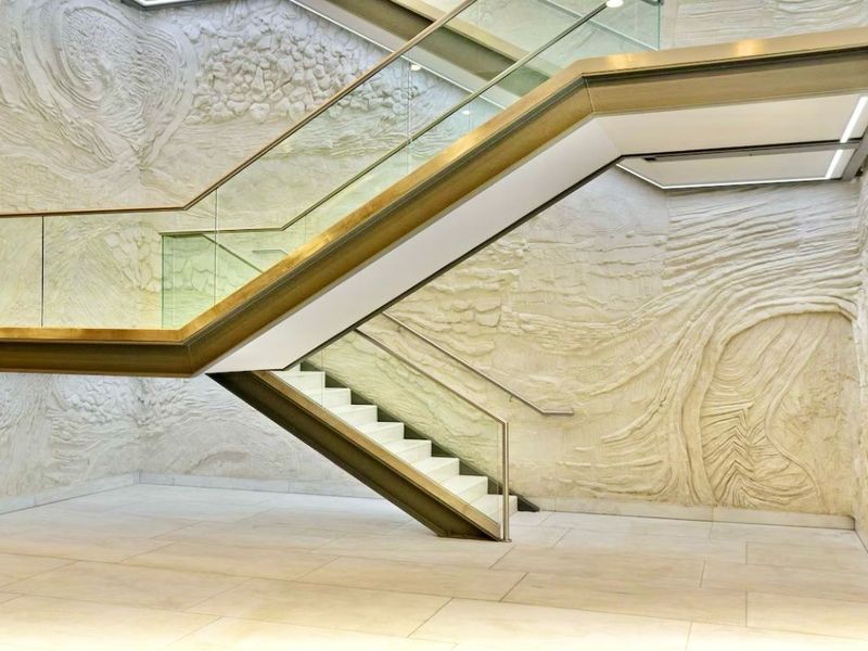 A modern architectural staircase featuring a glass balustrade, metal frame, and intricate textured feature wall in a spacious, high-end hallway.