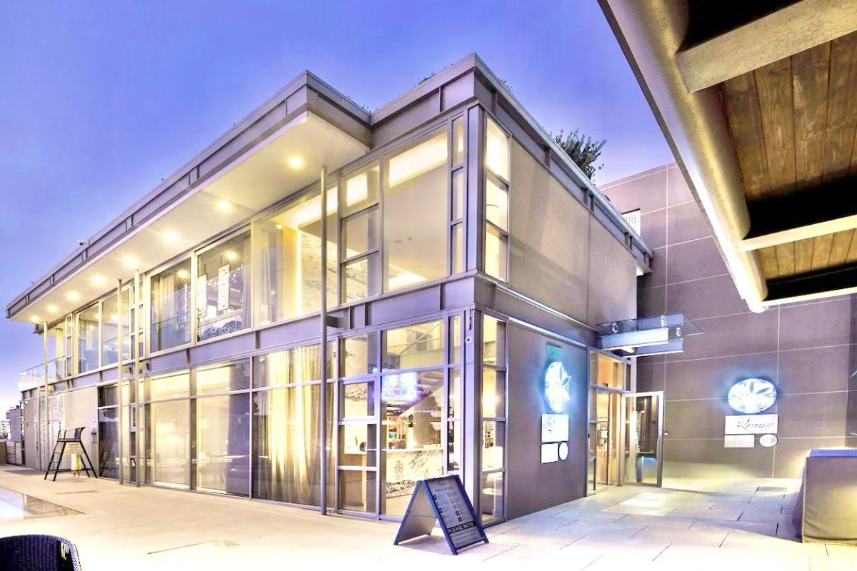 Modern multi-story apartment building exterior featuring extensive glass curtain walls, recessed lighting, and a paved terrace area at dusk.