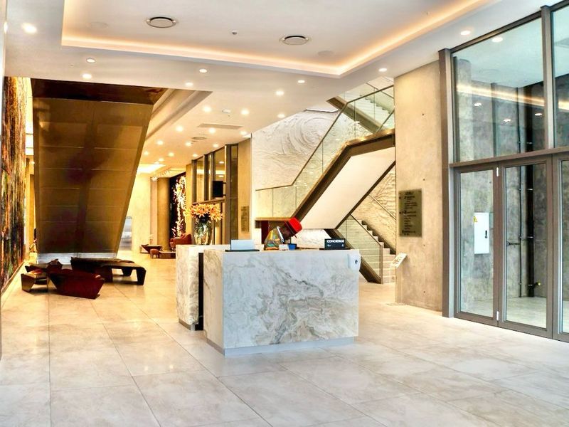 Modern apartment building lobby featuring a marble concierge desk, stone flooring, recessed lighting, and a striking glass-railed staircase.