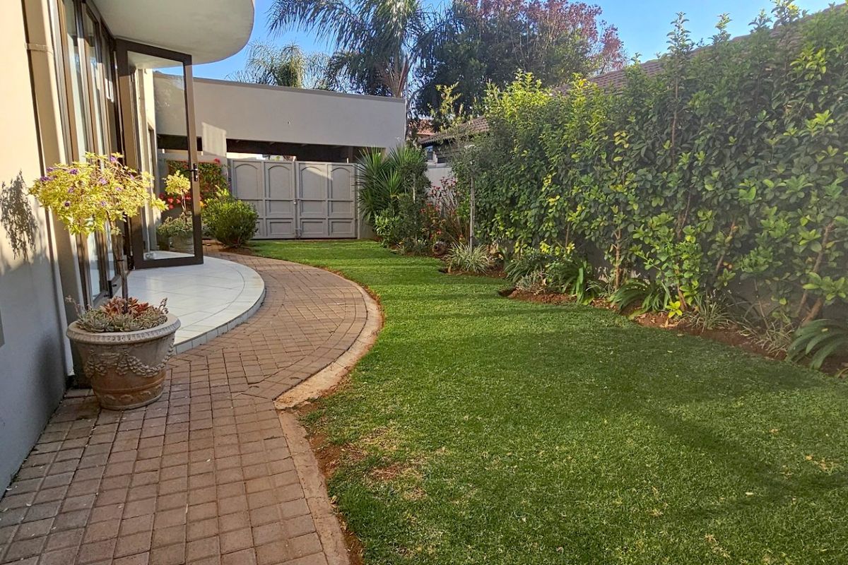 A well-maintained rear garden featuring a paved walkway, manicured lawn, and established boundary hedging, providing a private outdoor space.