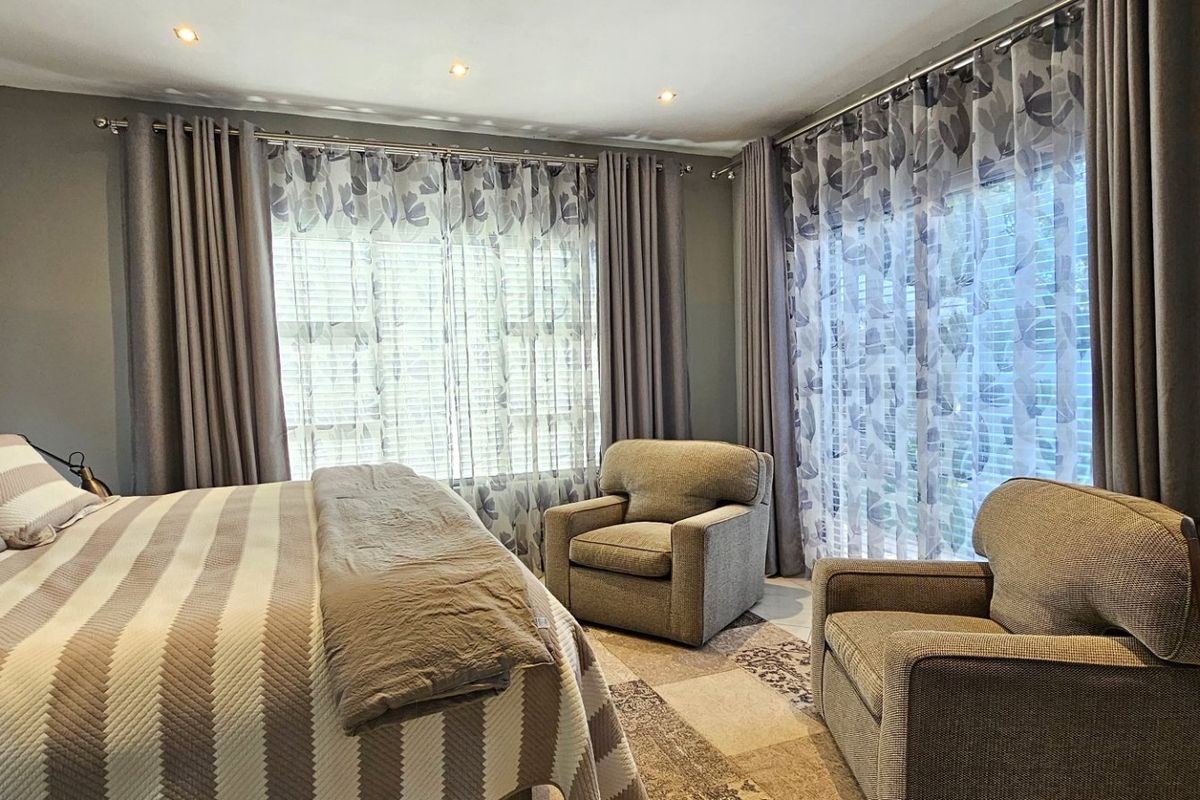 A well-maintained bedroom featuring neutral tones, recessed downlighting, and large windows dressed with layered curtains.