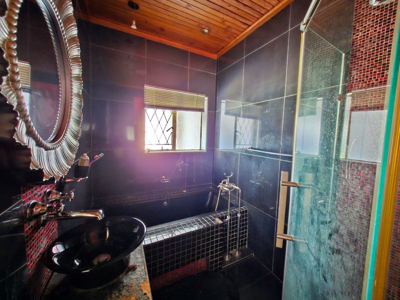 A compact bathroom featuring dark tiled walls, a built-in bathtub, a glass shower enclosure, and a timber ceiling.