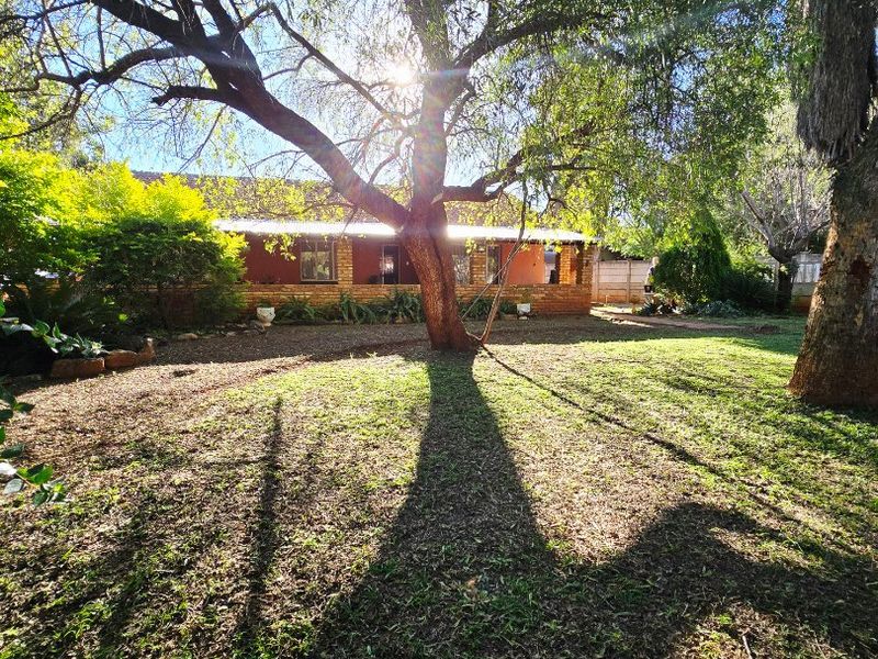 A spacious rear garden featuring mature trees and a view of the brick-built house with a covered patio area.