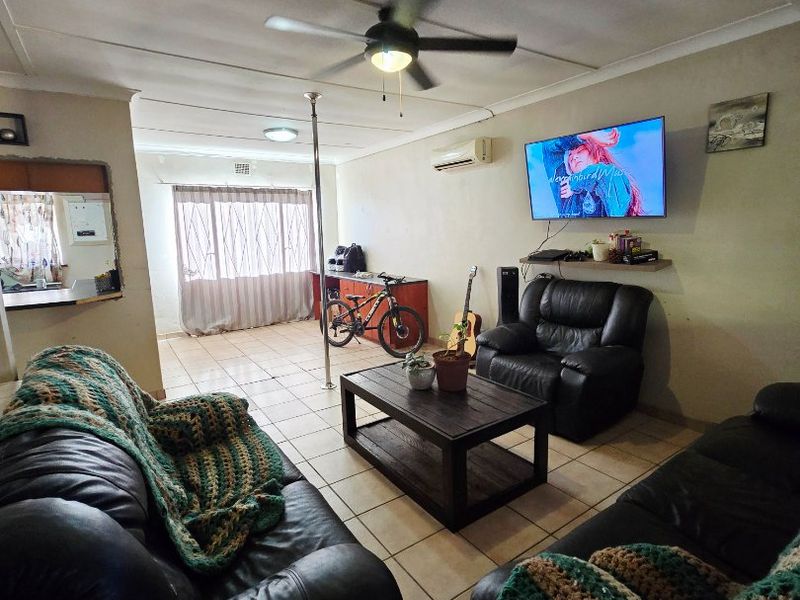 A living room featuring tiled flooring, a ceiling fan, and a wall-mounted air conditioning unit, with a pass-through window connecting to the kitchen area.
