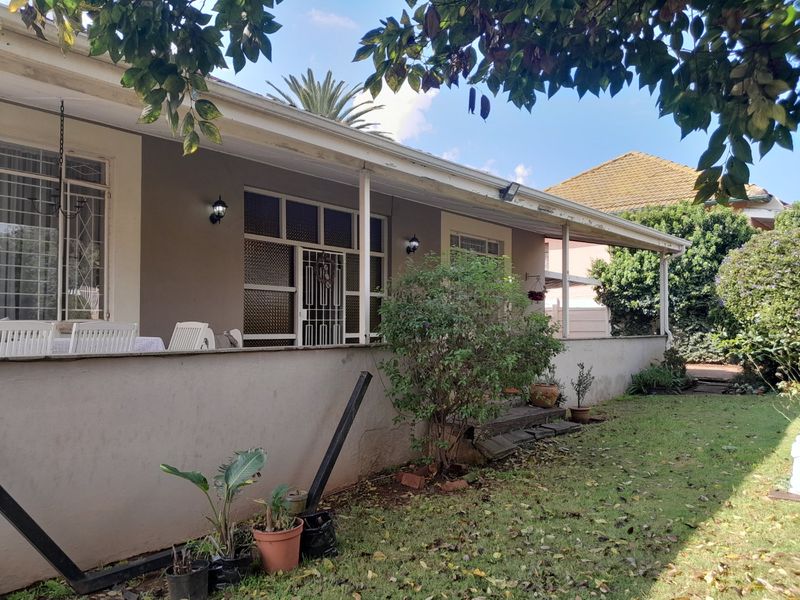 A rear exterior view of the house featuring a covered patio area with security gates and bars, showing signs of weathered exterior finishes.