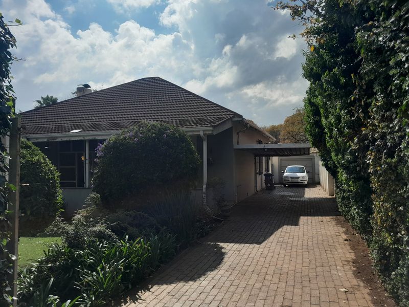 Front view of a single-story house featuring a paved driveway, carport, and garage, set within a mature garden.