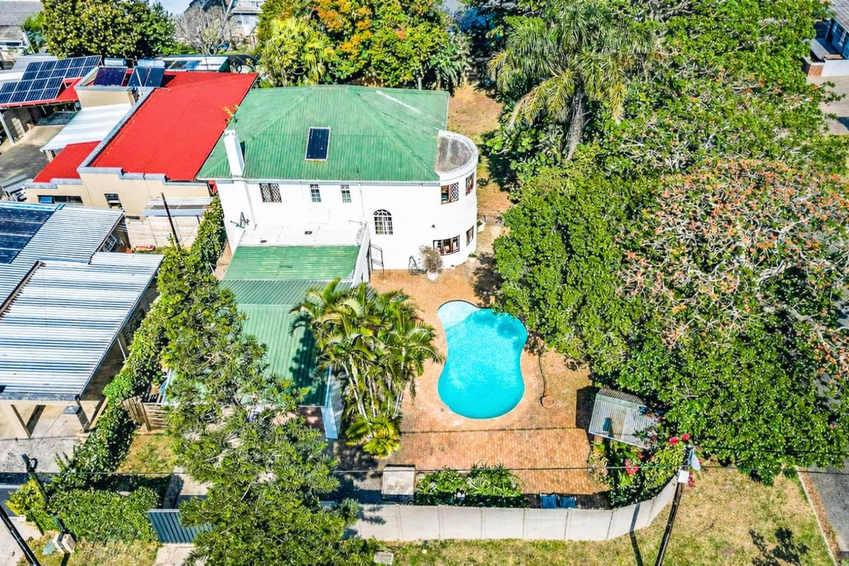 Aerial view of the property featuring a swimming pool, landscaped garden, brick paved patio area, and a multi-level house with a green metal roof.