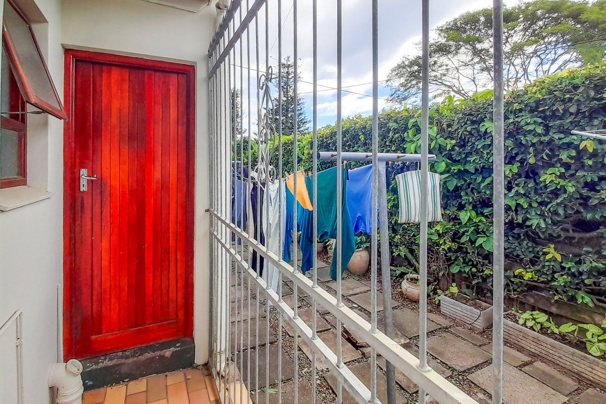 A rear exterior area featuring a paved patio, a secure metal gate, and a mature hedge boundary.